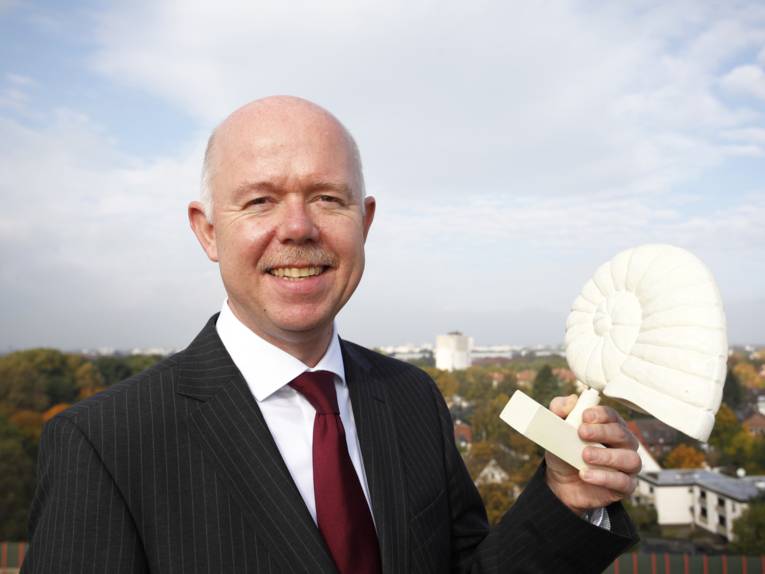 Ein Mann hält das Modell einer Schnecke in seiner linken Hand, im Hintergrund sind Gebäude und Bäume, der Betrachter erlebt den Hintergrund aus der Vogelperspektive.